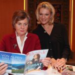 Zwei Frauen lesen an einem Tisch Zeitschriften; die eine sitzt und liest, die andere steht und lächelt neben ihr.