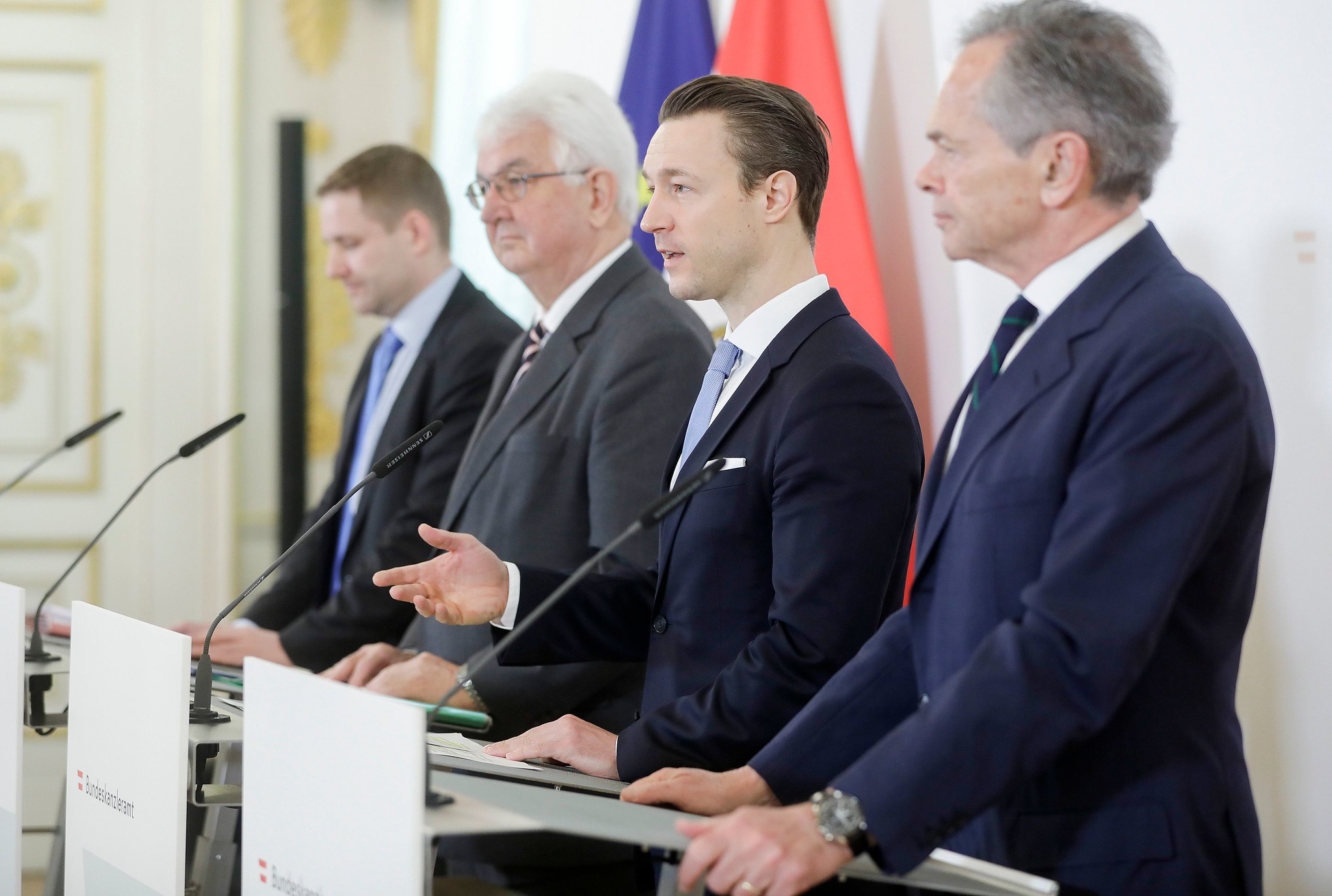 Vier Männer in Anzügen stehen auf einem Podium und sprechen auf einer formellen Pressekonferenz mit Fahnen im Hintergrund.