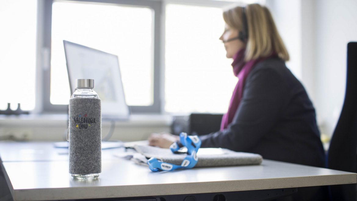 Eine gläserne Wasserflasche auf einem Schreibtisch mit einer Frau, die im Hintergrund an einem Computer arbeitet.