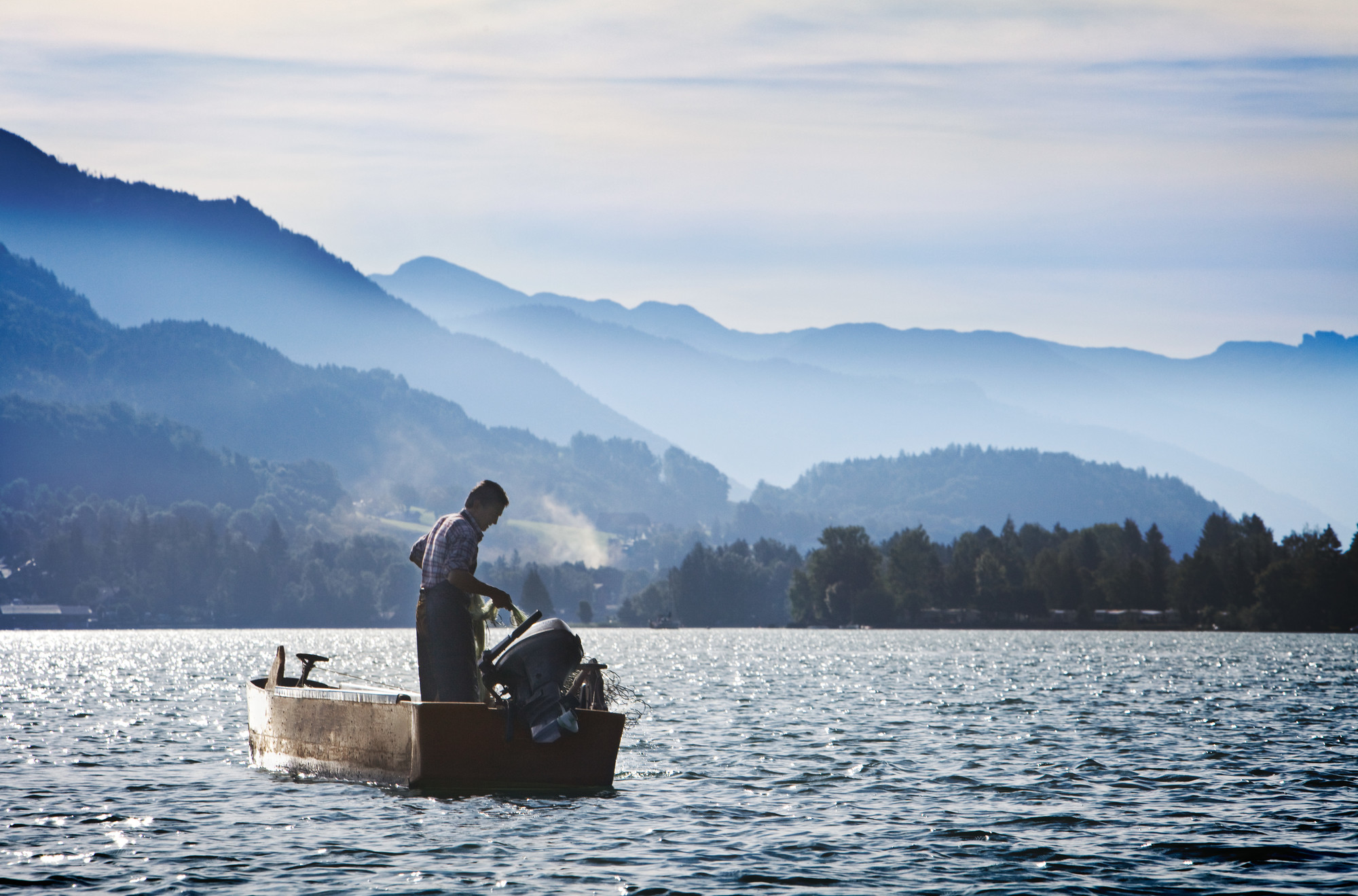 Eine Person steht in einem kleinen Boot auf einem See, umgeben von nebligen Bergen unter blauem Himmel.