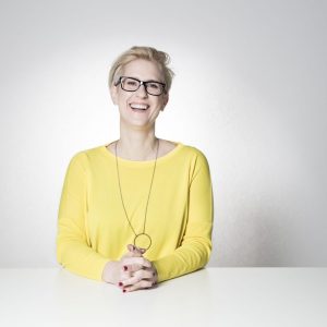 Lächelnde Frau mit kurzen blonden Haaren und Brille, die ein gelbes Oberteil trägt und an einem weißen Tisch sitzt.