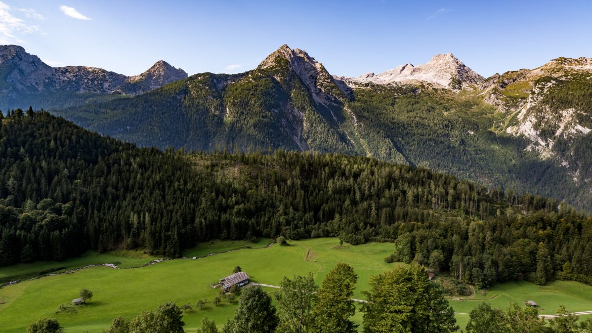 Ein grünes Tal mit verstreuten Hütten, dichten Kiefernwäldern und hohen Bergen unter einem klaren blauen Himmel.