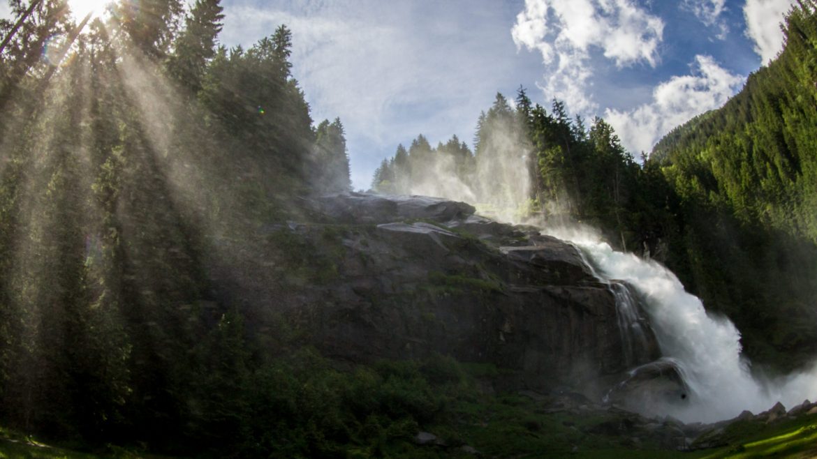 Ein Wasserfall ergießt sich in Kaskaden durch einen Wald, während die Sonnenstrahlen unter einem blauen Himmel durch hohe Kiefern scheinen.
