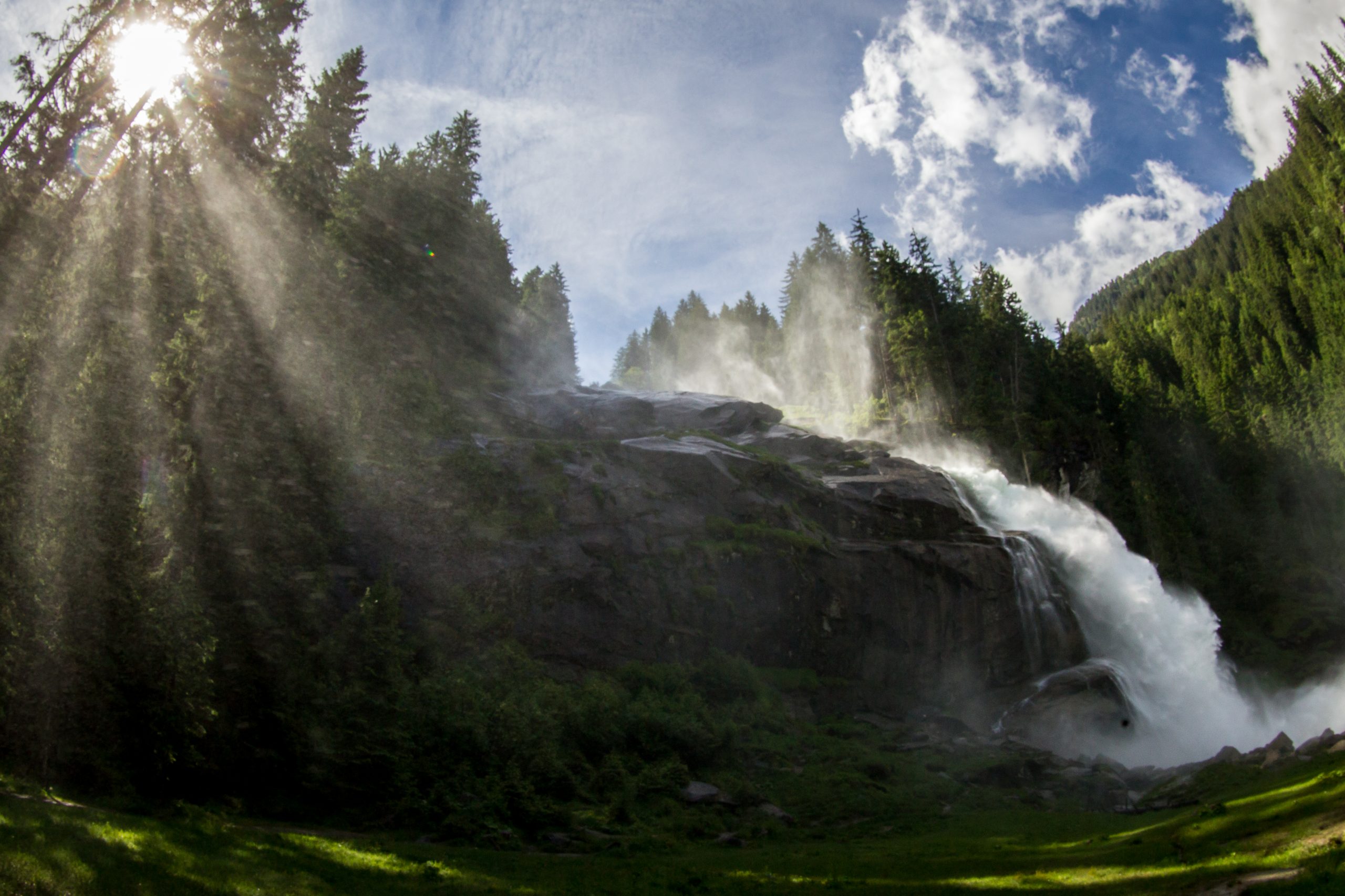 Ein Wasserfall ergießt sich in Kaskaden durch einen Wald, während die Sonnenstrahlen unter einem blauen Himmel durch hohe Kiefern scheinen.