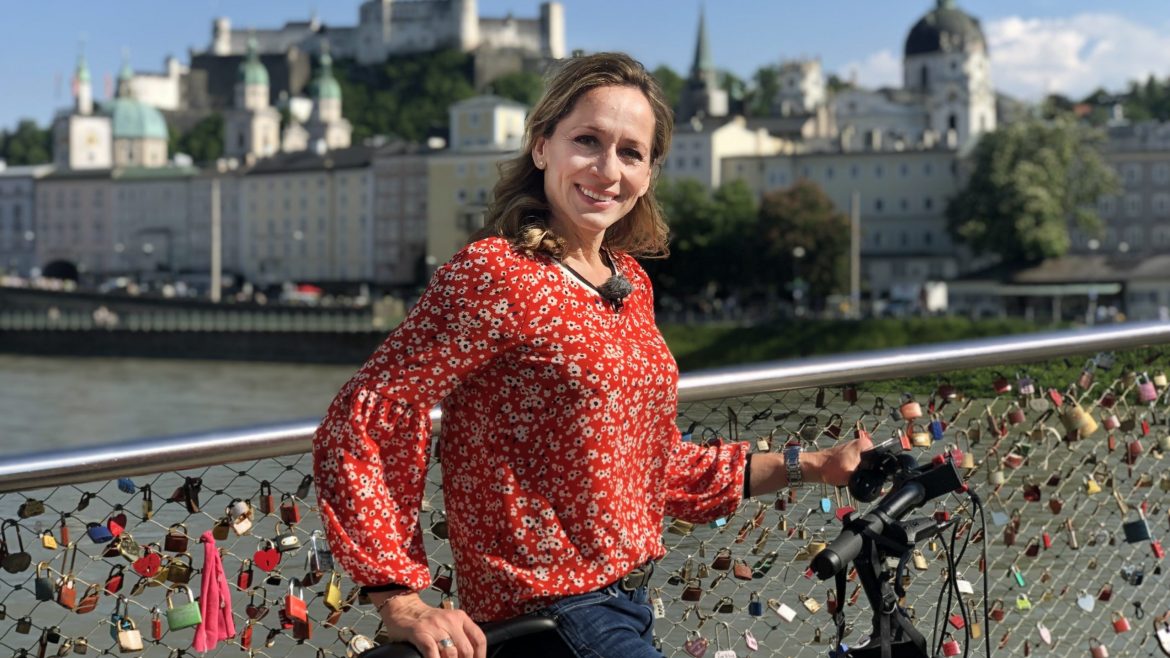 Frau in roter geblümter Bluse steht mit einem Fahrrad auf einer mit Liebesschlössern bedeckten Brücke, Stadtlandschaft im Hintergrund.