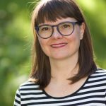 Frau mit braunem Haar und Brille lächelt, trägt ein schwarz-weiß gestreiftes Hemd, draußen im Sonnenlicht.