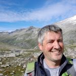 Ein lächelnder Mann macht ein Selfie in einer felsigen Berglandschaft mit blauem Himmel und Wolken.