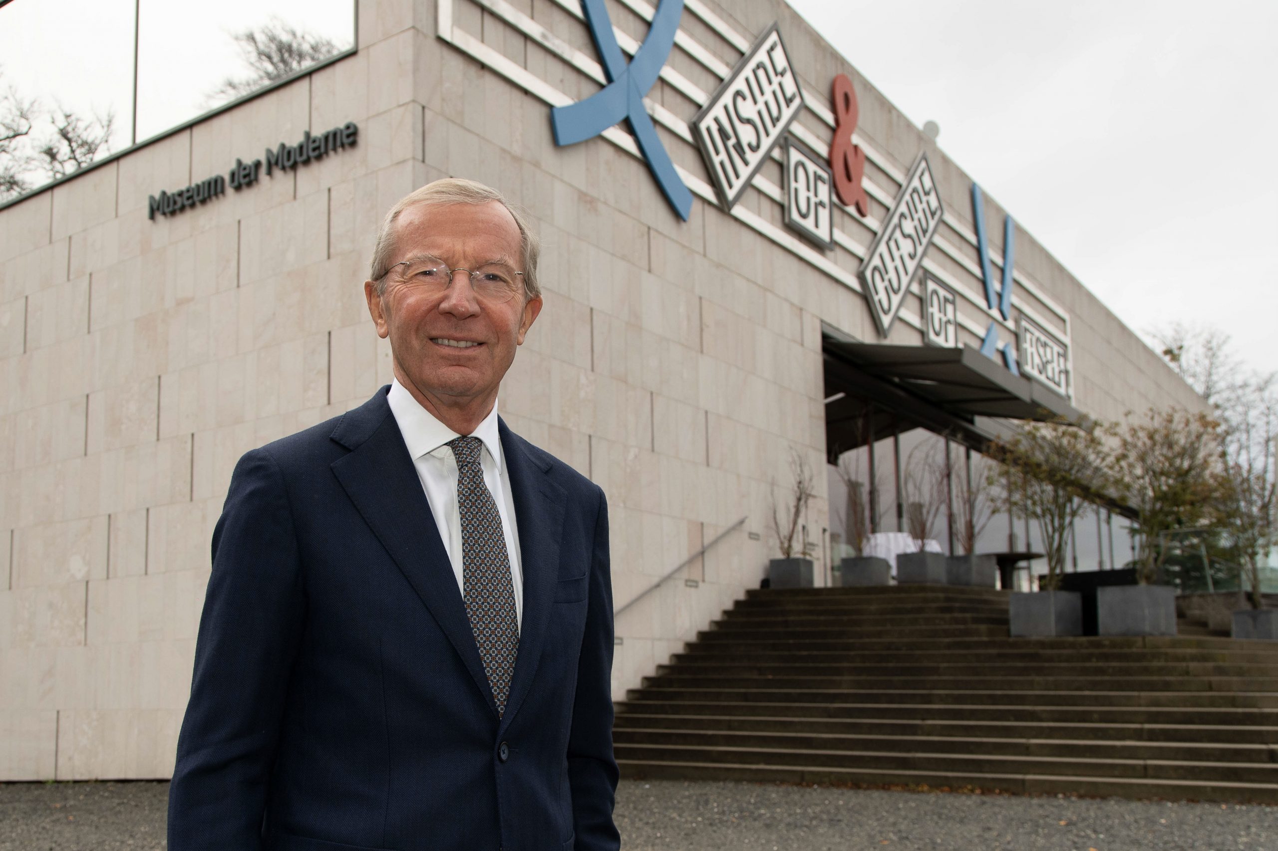 Ein lächelnder Mann im Anzug steht vor dem Gebäude des Museums der Moderne mit einer Beschilderung für moderne Kunst darüber.