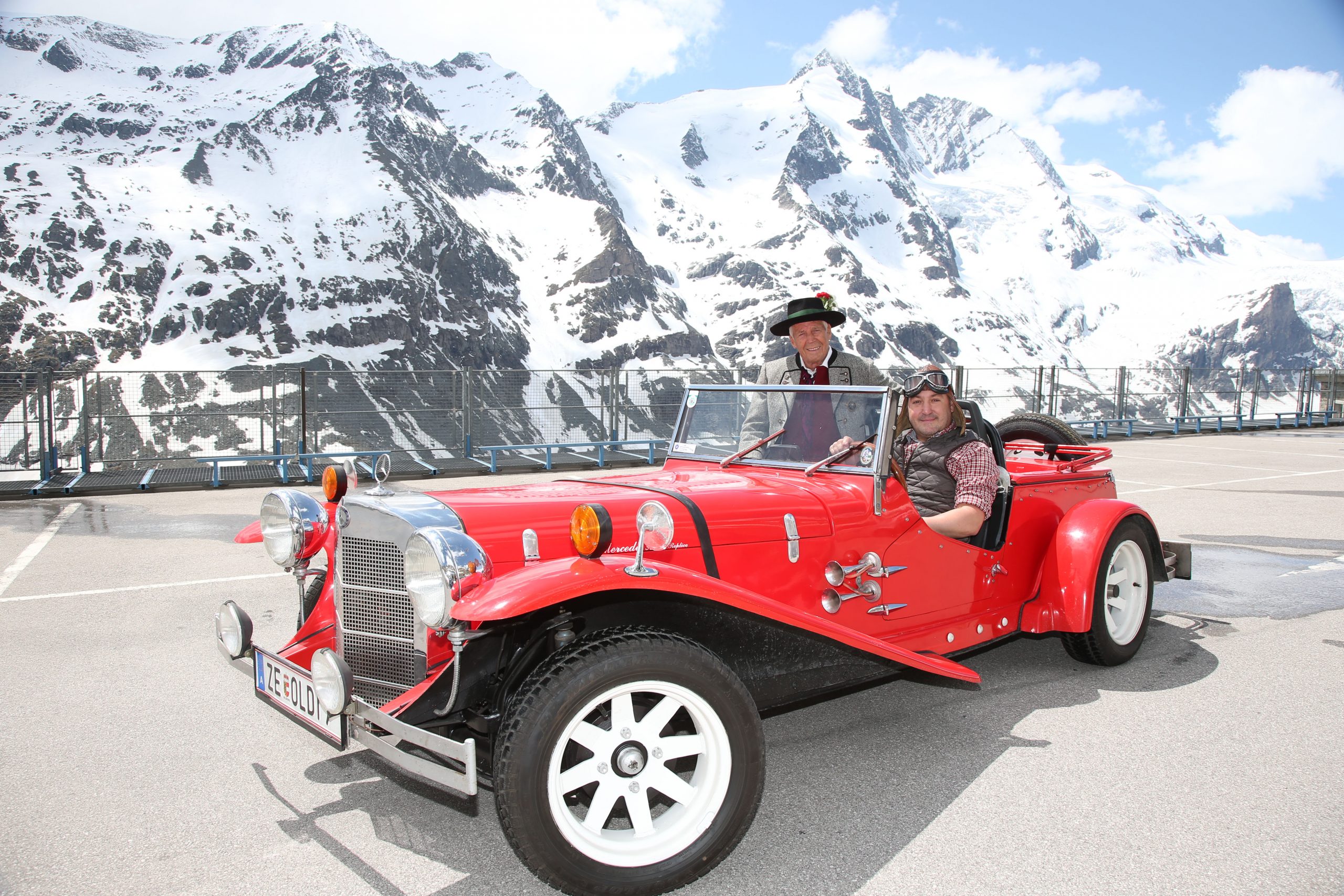 Zwei Personen in einem roten Oldtimer-Cabrio mit verschneiten Bergen im Hintergrund an einem sonnigen Tag.