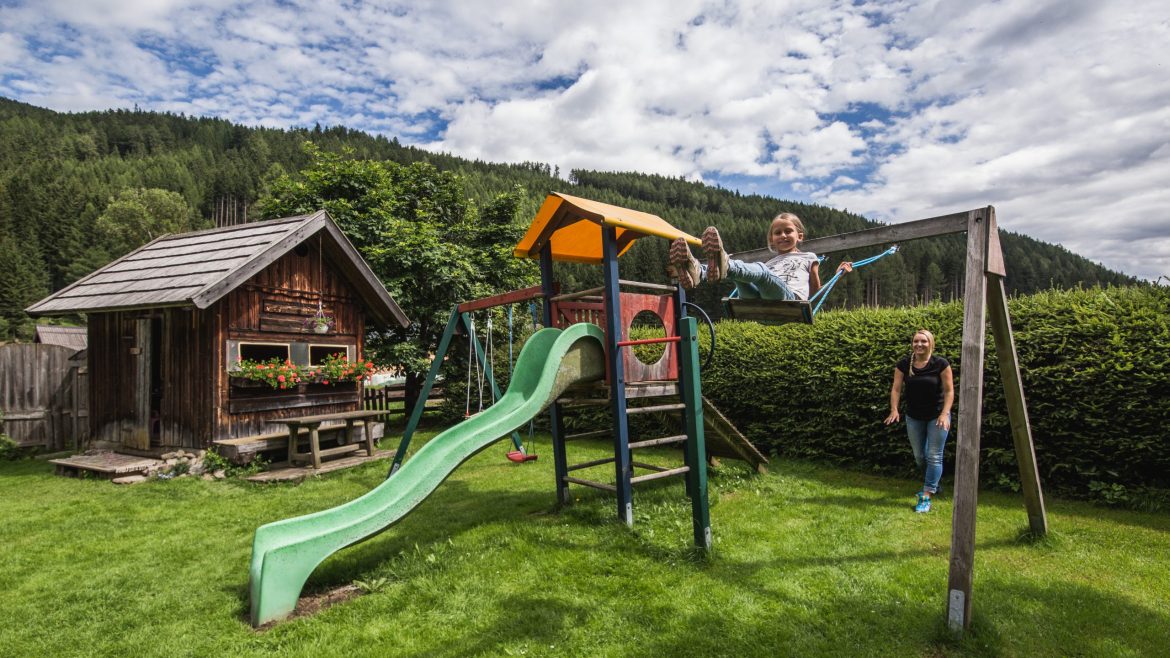 Ein Kind schaukelt neben einer Rutsche in einem grasbewachsenen Hof, im Hintergrund sind eine Frau und ein Holzhaus zu sehen.
