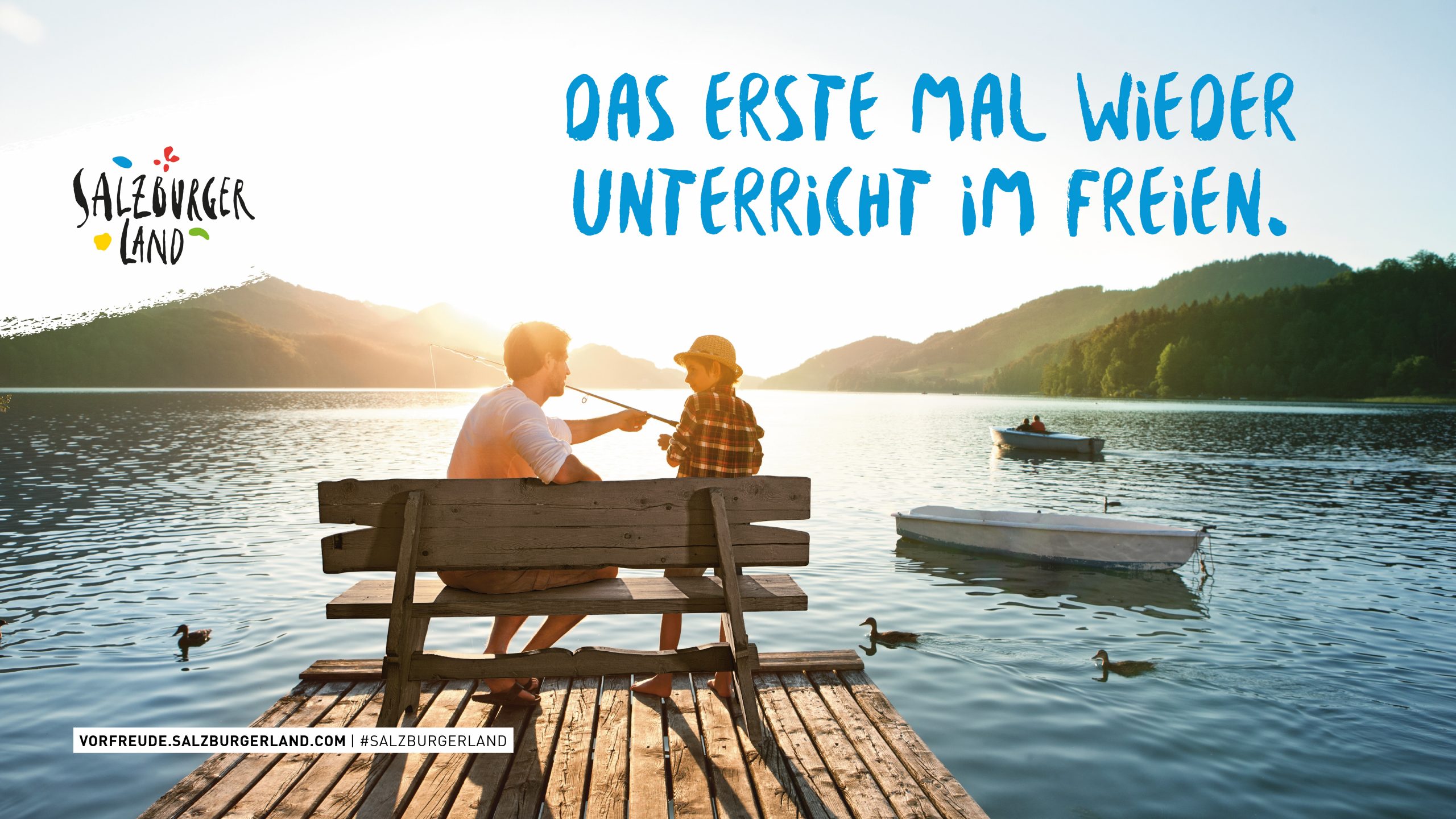 Zwei Personen sitzen auf einer Bank am Seeufer und angeln bei Sonnenuntergang mit Bergen und Wasser im Hintergrund.