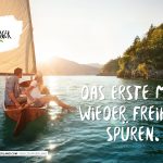 Ein Paar segelt bei Sonnenuntergang auf einem See, umgeben von Bergen; der Text lautet "Das erste Mal wieder Freiheit spüren.".