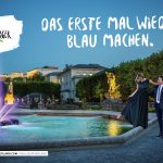 Ein elegant gekleidetes Paar spaziert bei Sonnenuntergang an einem farbenfrohen Springbrunnen in einem Park vorbei, darüber ein Text in deutscher Sprache.