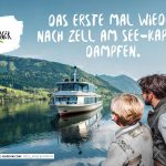 Zwei Personen in österreichischer Tracht schauen auf ein Boot auf einem See mit Bergen im Hintergrund.