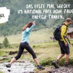 Zwei Wanderer überqueren einen felsigen Bach in einer grünen Berglandschaft in der Nähe einer Holzhütte im Nationalpark Hohe Tauern.