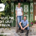 Zwei Personen lächeln vor einem rustikalen Gasthaus mit Schildern und Blumen, das Gäste in Salzburg, Österreich, willkommen heißt.