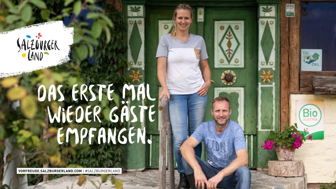 Ein lächelnder Mann und eine lächelnde Frau stehen vor einer rustikalen Holztür mit dem Text Das erste Mal wieder Gäste empfangen.