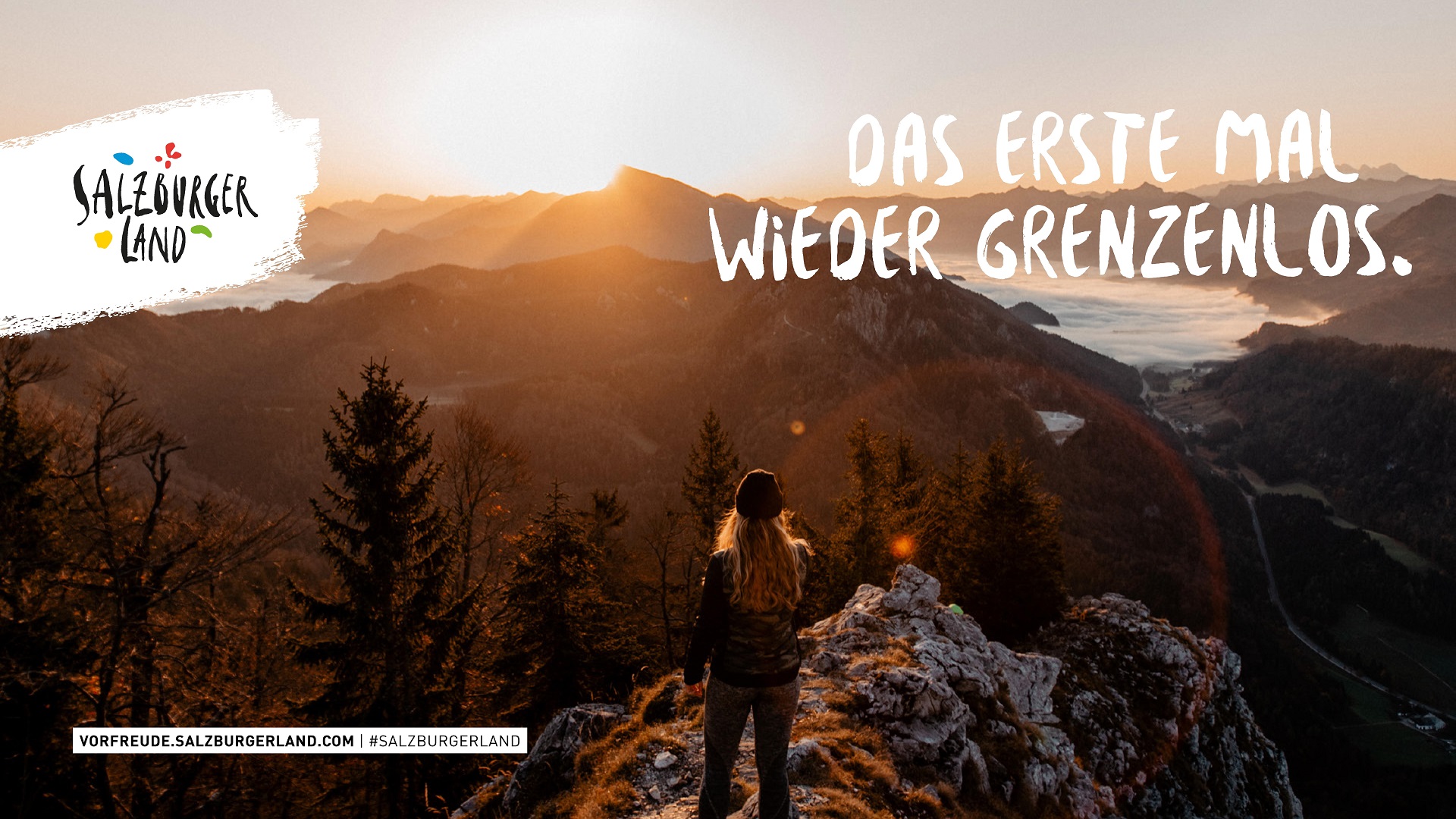 Eine Person steht bei Sonnenaufgang auf einem Berggipfel und blickt auf ein malerisches Tal und ferne Berge.