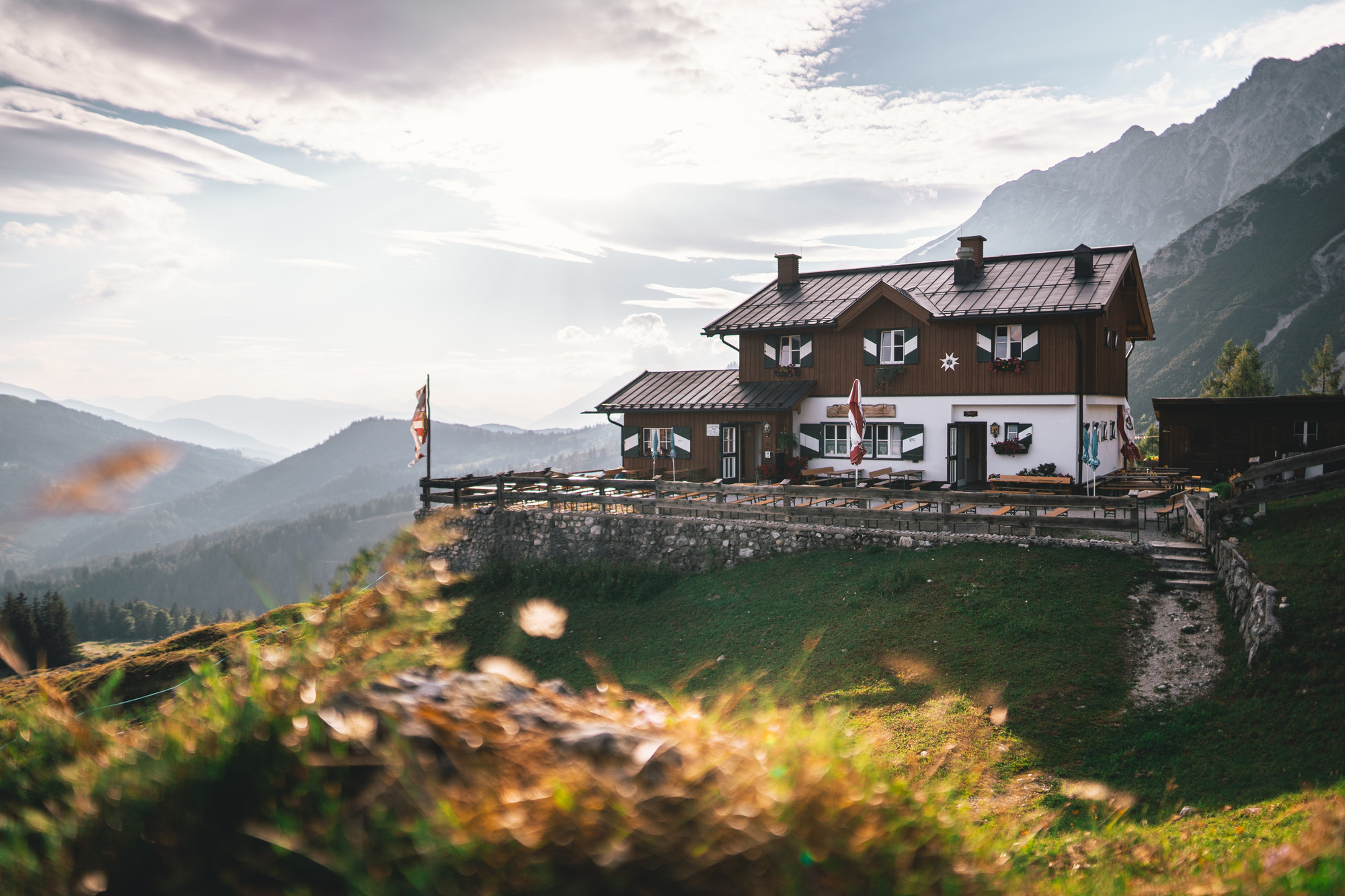 Eine hölzerne Berghütte liegt auf einer grasbewachsenen Anhöhe mit malerischen Bergen und einem strahlenden Himmel im Hintergrund.