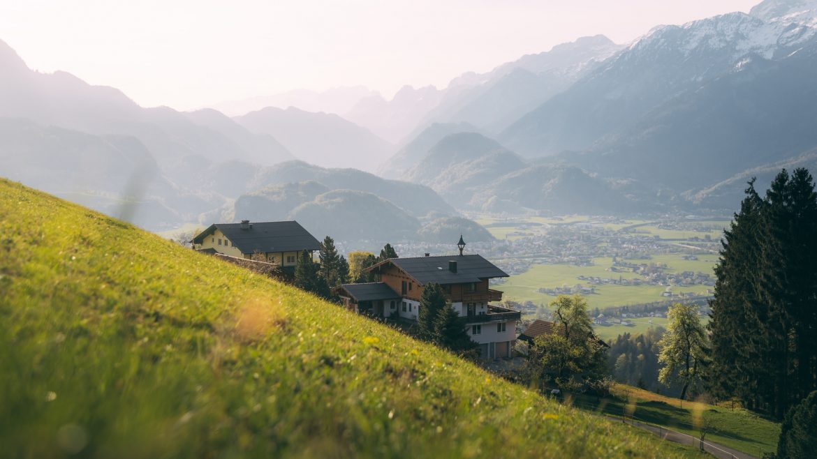 Häuser auf einem grasbewachsenen Hügel überblicken ein grünes Tal mit Bergen im Hintergrund unter einem dunstigen Himmel.