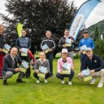 Eine Gruppe von neun Personen, die im Freien auf dem Rasen sitzen, Zeitschriften in der Hand halten und in die Kamera lächeln, neben Bannern von Golfplätzen.