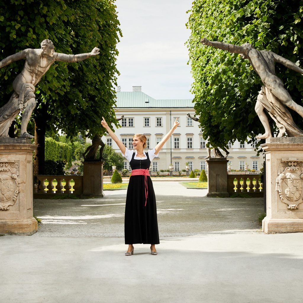 Frau in traditioneller Kleidung steht zwischen zwei Statuen mit erhobenen Armen in einem Garten mit Bäumen und einem großen Gebäude.