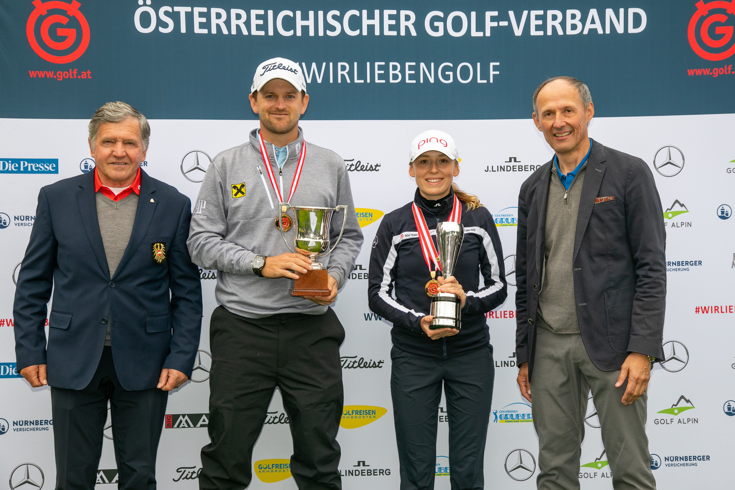 Vier Personen posieren bei einer Golfveranstaltung; zwei von ihnen halten Trophäen und Medaillen und stehen vor einem Markenhintergrund.