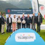 Eine Gruppe von Personen posiert bei einer Golfveranstaltung mit Trophäen und Bannern vor Bergen.