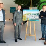 Drei Männer stehen neben einer Staffelei, auf der ein Stadtfoto in einem hellen Innenraum zu sehen ist.