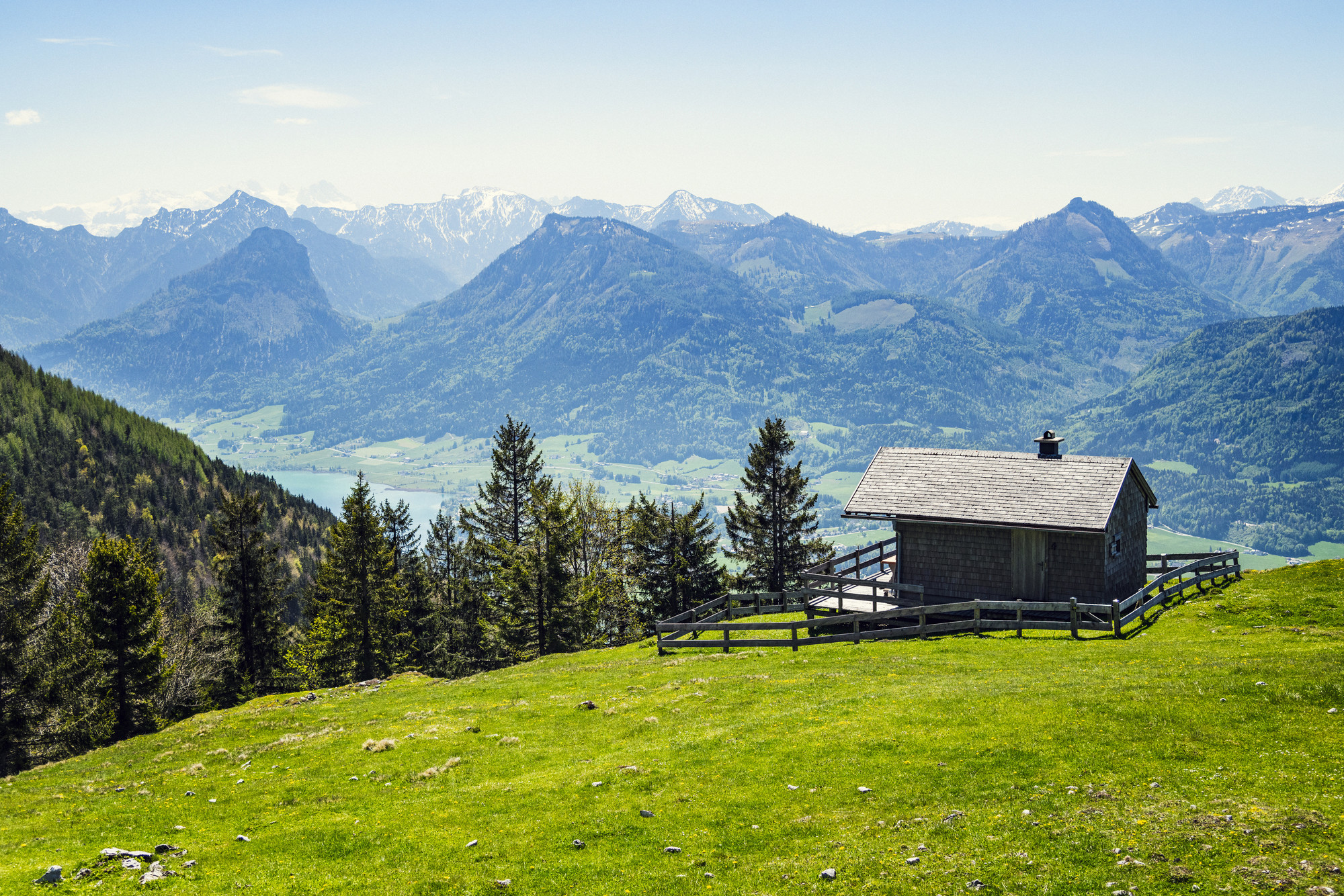 Eine kleine Hütte auf einem grasbewachsenen Hügel überblickt die fernen Berge und einen See unter einem klaren blauen Himmel.