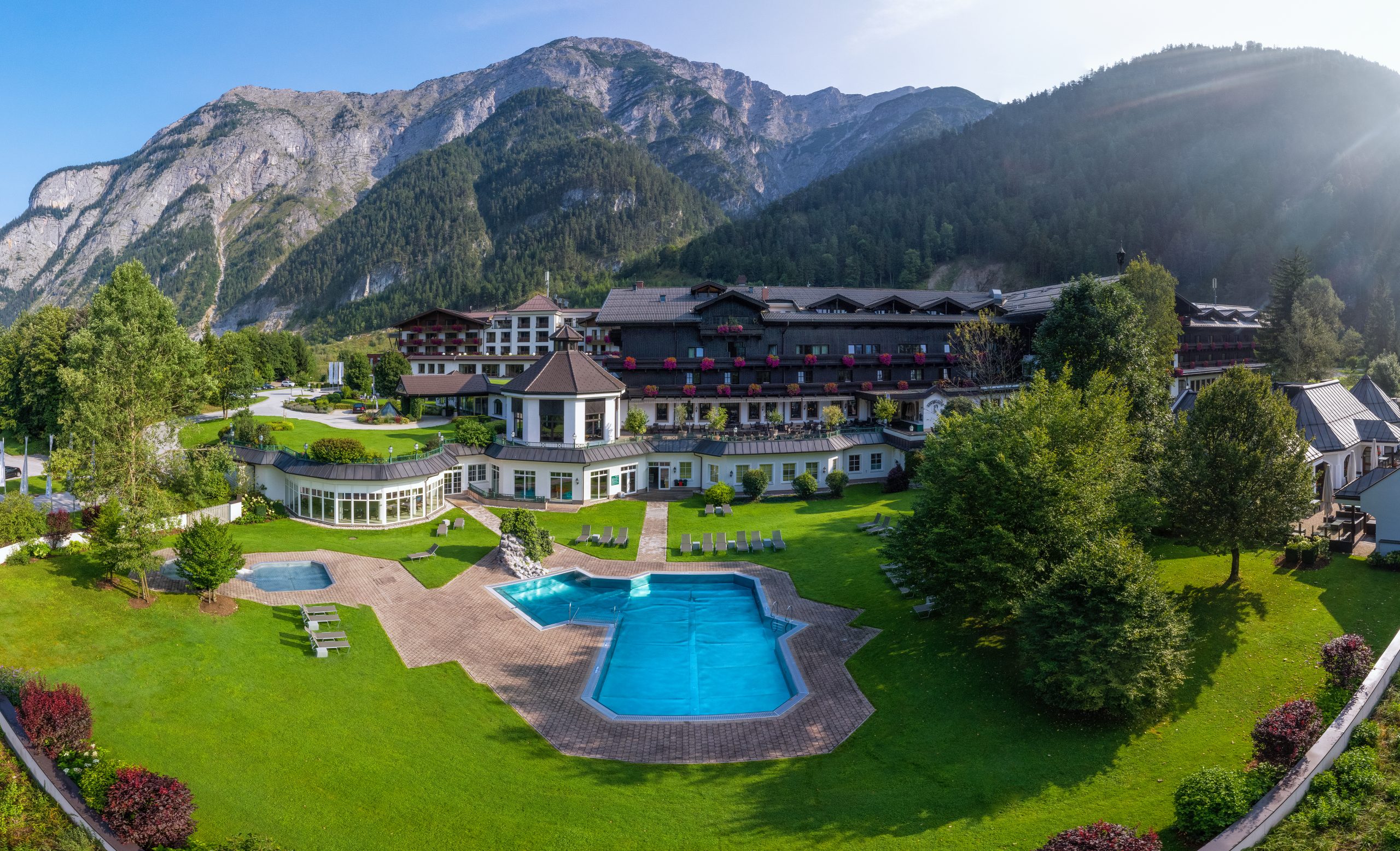Ein Luxushotel mit Pool und Gärten, umgeben von bewaldeten Bergen und einem klaren Himmel.