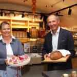 Zwei Personen lächeln im Haus, die eine hält ein Tablett mit Gebäck, die andere einen Laib Brot auf einem Brett.