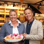 Zwei Personen lächeln und halten einen Teller mit bunten Cupcakes in einer Bäckerei oder einem Café.