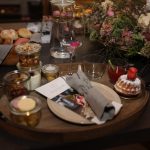 Ein Holztablett mit Desserts, Nüssen, Getränken und einer Serviette steht auf einem Tisch mit Blumen und Brötchen.