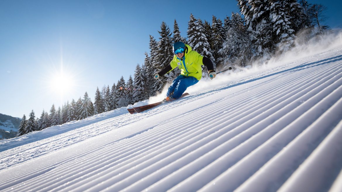 Ein Skifahrer in heller Ausrüstung fährt eine präparierte Schneepiste mit Bäumen und Sonnenschein im Hintergrund hinunter.