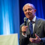 Ein älterer Mann im Anzug spricht in ein Mikrofon auf der Bühne mit blauer Beleuchtung im Hintergrund.