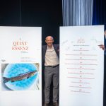 Drei Männer in Anzügen und Kochkleidung stehen neben zwei großen, signierten Plakaten auf einer Veranstaltungsbühne.