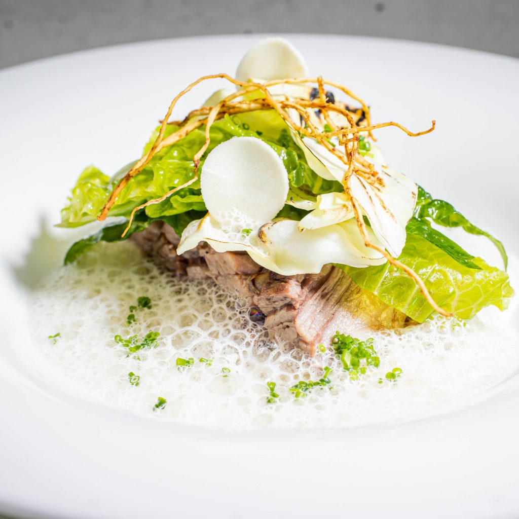 Ein Gourmet-Gericht mit Grünzeug, Schaum, dünn geschnittenem Gemüse und Beilagen auf einem weißen Teller.