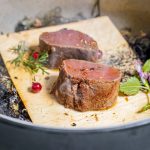 Zwei Stücke gebratenes Steak auf einem Holzbrett, garniert mit Kräutern und Beeren, umgeben von rauchigen Elementen.