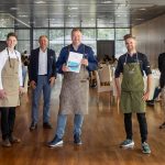 Sieben Männer stehen in einem hellen Speisesaal und posieren für ein Foto, einige tragen Schürzen, einer hält eine Urkunde.