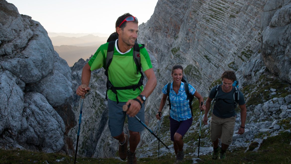Drei Personen wandern mit Trekkingstöcken einen felsigen Bergpfad hinauf, umgeben von steilen Felswänden und fernen Gipfeln.