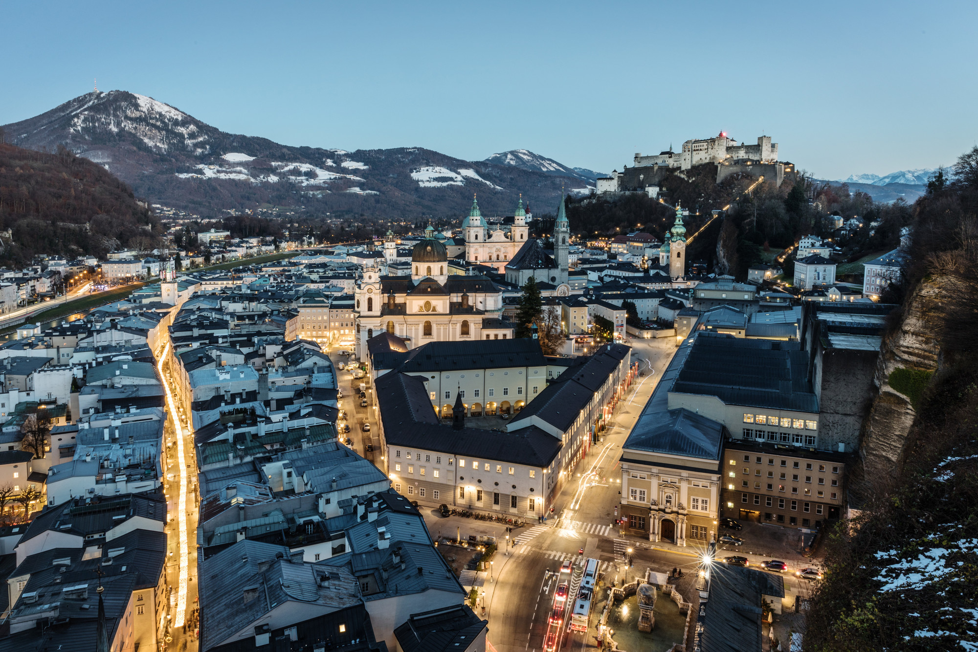Luftaufnahme von Salzburg in der Abenddämmerung, mit beleuchteten Straßen, historischen Gebäuden und einer Festung auf einem Hügel.