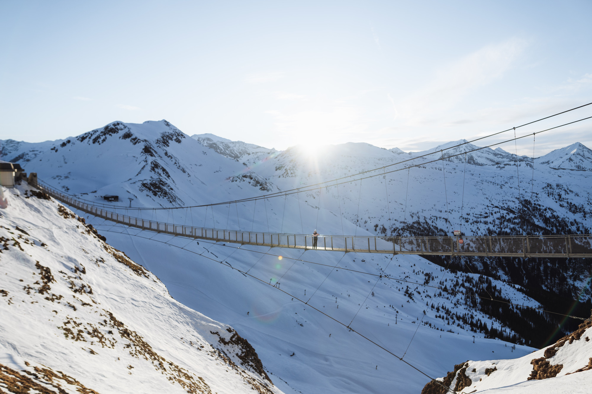 Eine Hängebrücke überquert bei Sonnenaufgang verschneite Berge, in deren Mitte eine Person läuft.