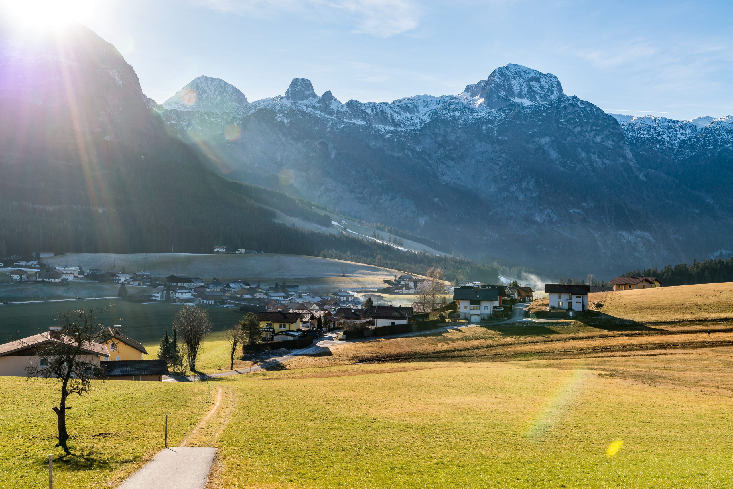 Ein sonnenbeschienenes Alpendorf mit Bergen im Hintergrund und grünen Feldern im Vordergund.