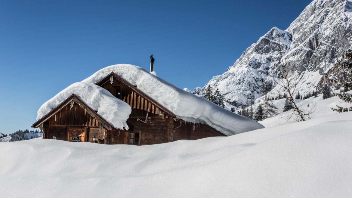 Eine schneebedeckte Holzhütte steht vor verschneiten Bergen unter einem strahlend blauen Himmel.