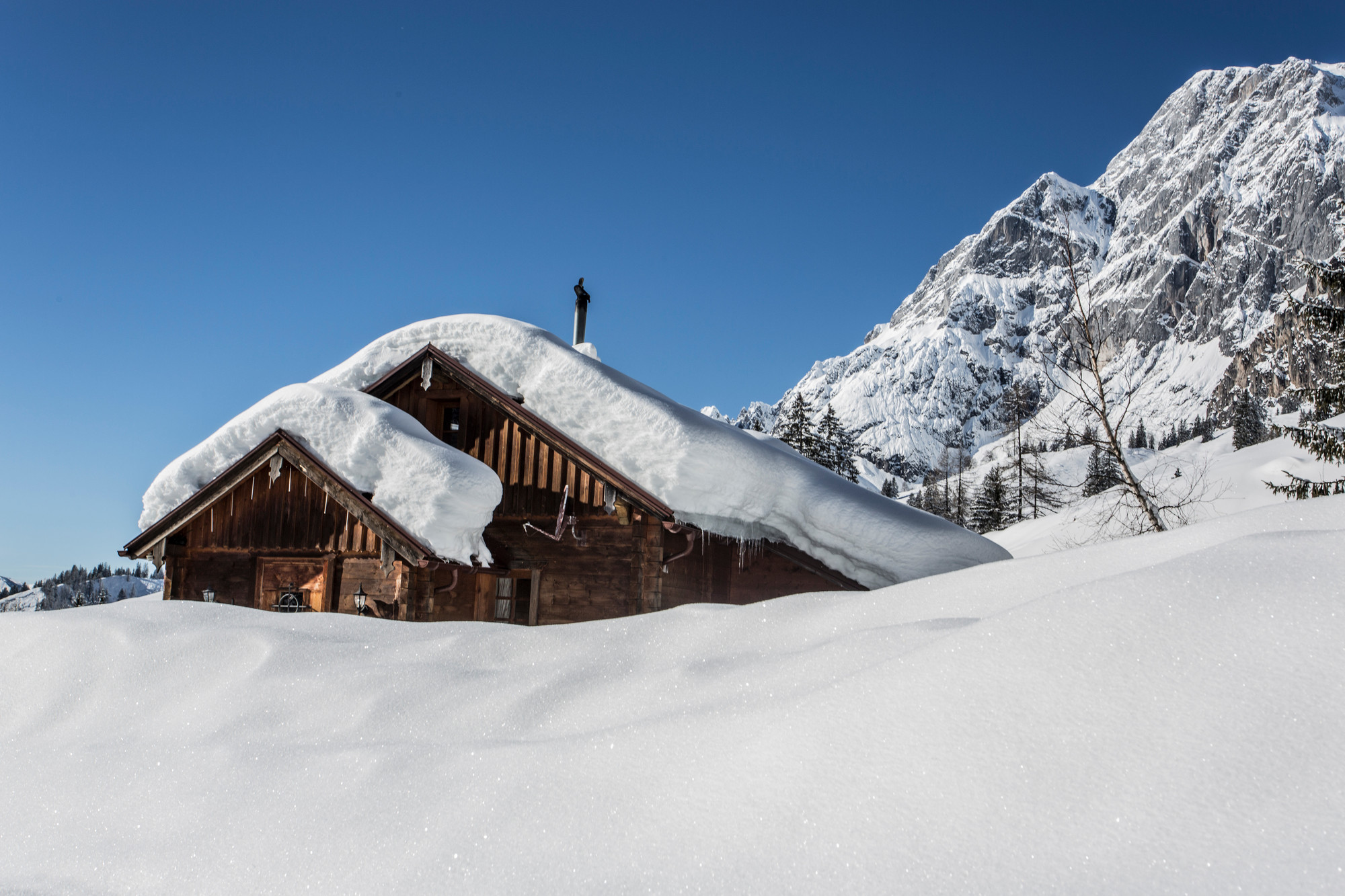 Eine schneebedeckte Holzhütte steht vor verschneiten Bergen unter einem strahlend blauen Himmel.