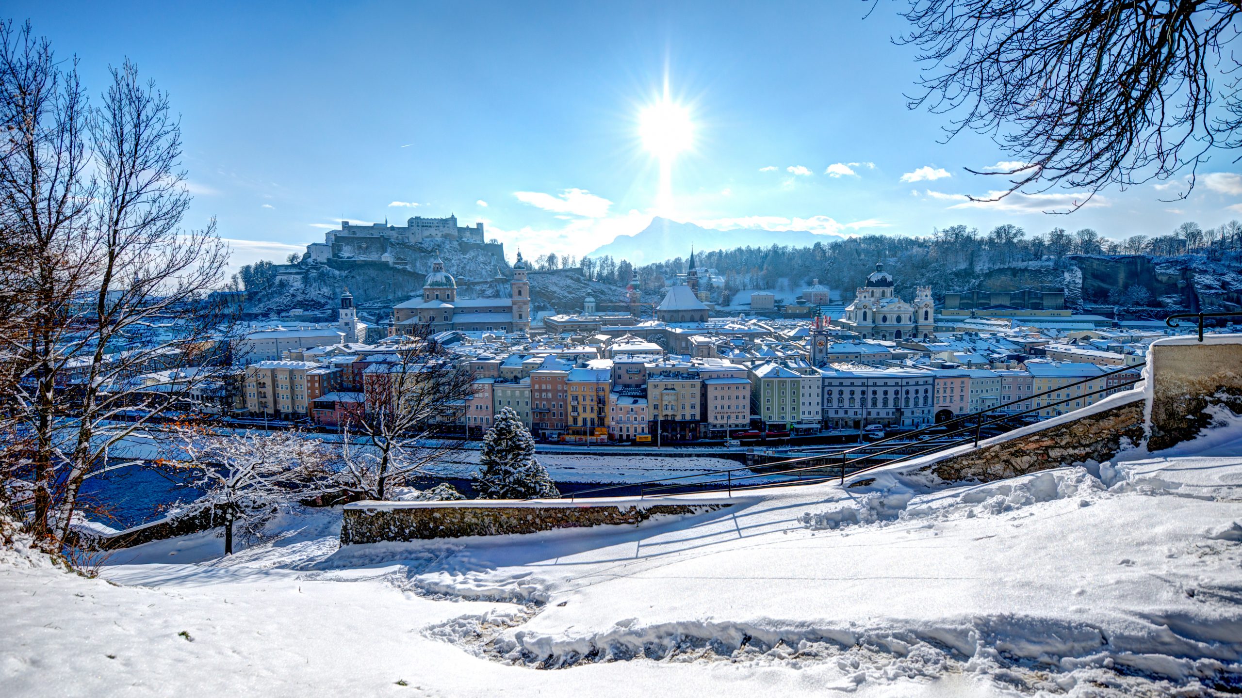 Schneebedeckte Stadtlandschaft von Salzburg, Österreich, mit Sonnenschein und einer Festung auf einem entfernten Hügel.