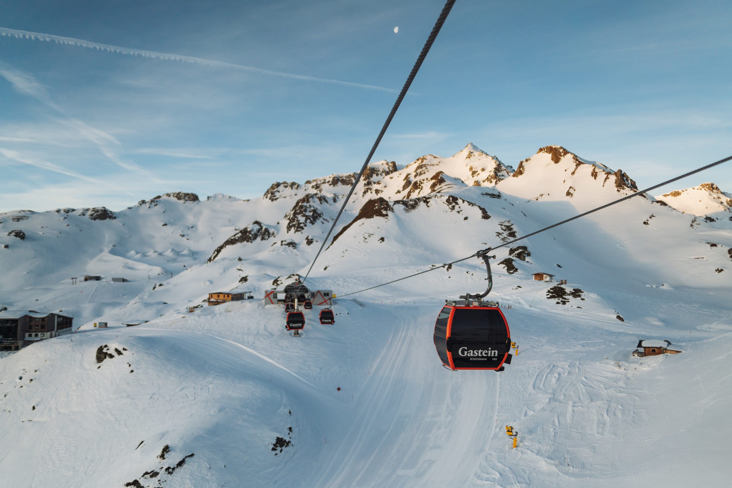Rote Seilbahnen fahren über verschneite Berghänge unter einem klaren Himmel mit vereinzelten Wolken und weit entfernten Gipfeln.