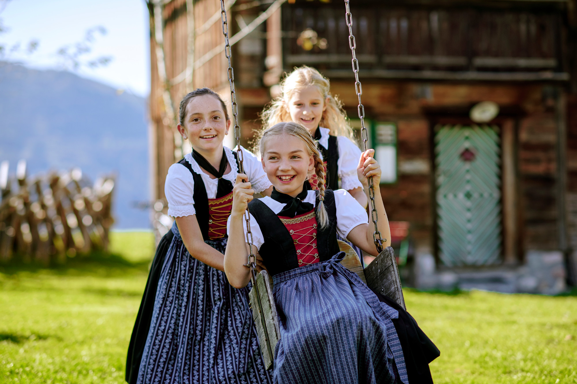 Drei lächelnde Mädchen in österreichischen Trachten, die an einem sonnigen Tag draußen in der Nähe eines rustikalen Holzhauses schaukeln und spielen.