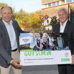 Zwei Männer in Anzügen halten ein großes Gutscheinschild, auf dem lächelnde Mädchen in österreichischer Tracht abgebildet sind.